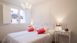 A bedroom with two beds, a chandelier, a bedside table with a lamp, and a window with shutters.