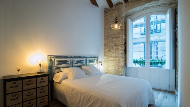 A bedroom with a bed, a dresser with drawers, a hanging light fixture, and a window with a view of greenery. - PPRINCIPAL D - PD - APARTAMENTOS ALMADA - Plaza del Pilar (Zaragoza)