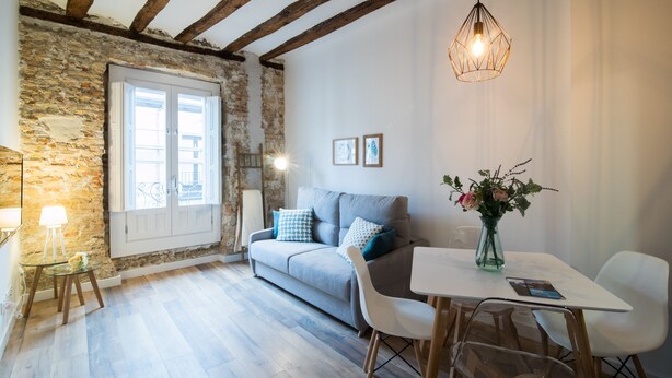 A cozy room with a stone wall, a wooden ceiling, a grey sofa, a white table, and a hanging light fixture. - SEGUNDO D - 2D - APARTAMENTOS ALMADA - Plaza del Pilar (Zaragoza)