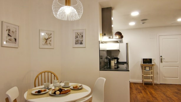 A dining area with a round table, white chairs, and a modern pendant light. There are framed pictures on the wall, a microwave on a small shelf, and a door leading to another room. - ENTRESUELO A - E A - Apartamentos Almada - Plaza del Pilar (Zaragoza)
