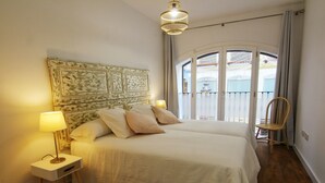 A bedroom with a large bed, two bedside tables with lamps, a wooden chair, and a balcony with a view. - ENTRESUELO A - E A - Apartamentos Almada - Plaza del Pilar (Zaragoza)
