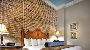 A hotel room with a brick wall, a wooden headboard, a bed with white linens, a blue throw, and a wooden nightstand with a lamp.