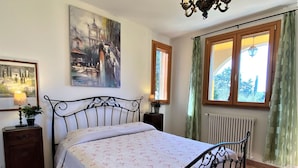 Un dormitorio con cama, mesita de noche, lámpara, un cuadro, ventana con cortinas y radiador.