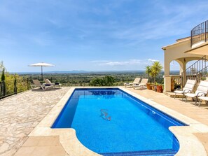 Une piscine rectangulaire à l’eau d’un bleu limpide, entourée de carreaux de pierre et d’une terrasse en pierre. On y trouve des chaises longues et un parasol blanc.
