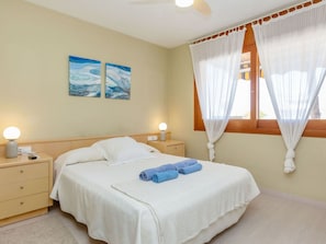 A bedroom with a bed, two bedside tables, a ceiling fan, a window with curtains, and two wall-mounted paintings.