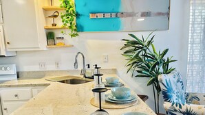 A kitchen with a granite countertop, a stainless steel sink, and a modern faucet. There are decorative plants and a large window with a view of a beach. - Surfside Serenity at Bidawee PCB (Panama City Beach)