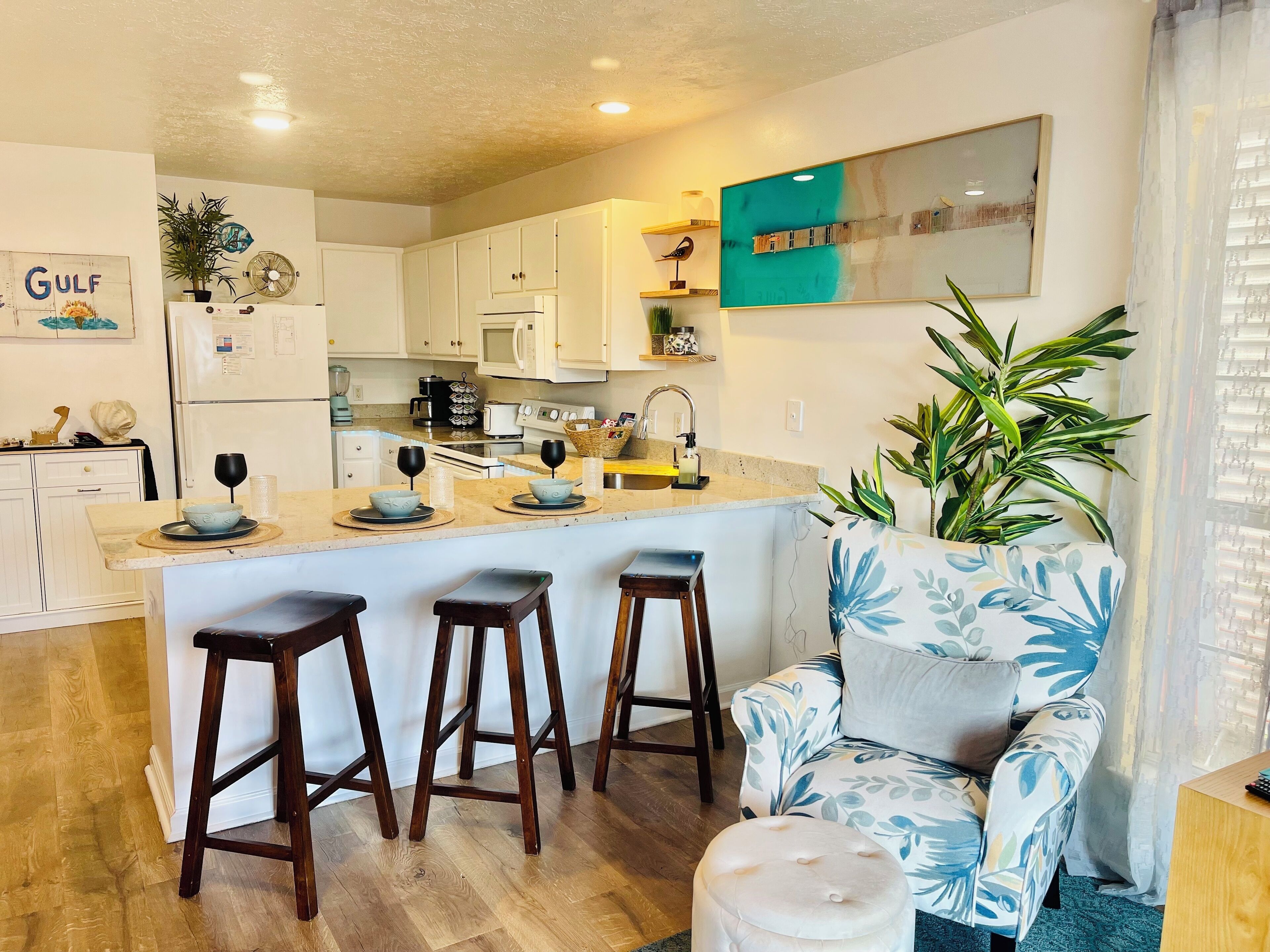 A modern kitchen with white cabinets, a central island with stools, a large potted plant, and a blue and white floral armchair.
