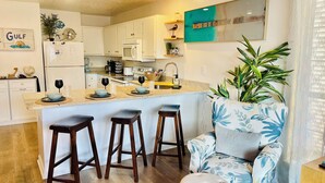 A modern kitchen with white cabinets, a central island with stools, a large potted plant, and a blue and white floral armchair. - Surfside Serenity at Bidawee PCB (Panama City Beach)