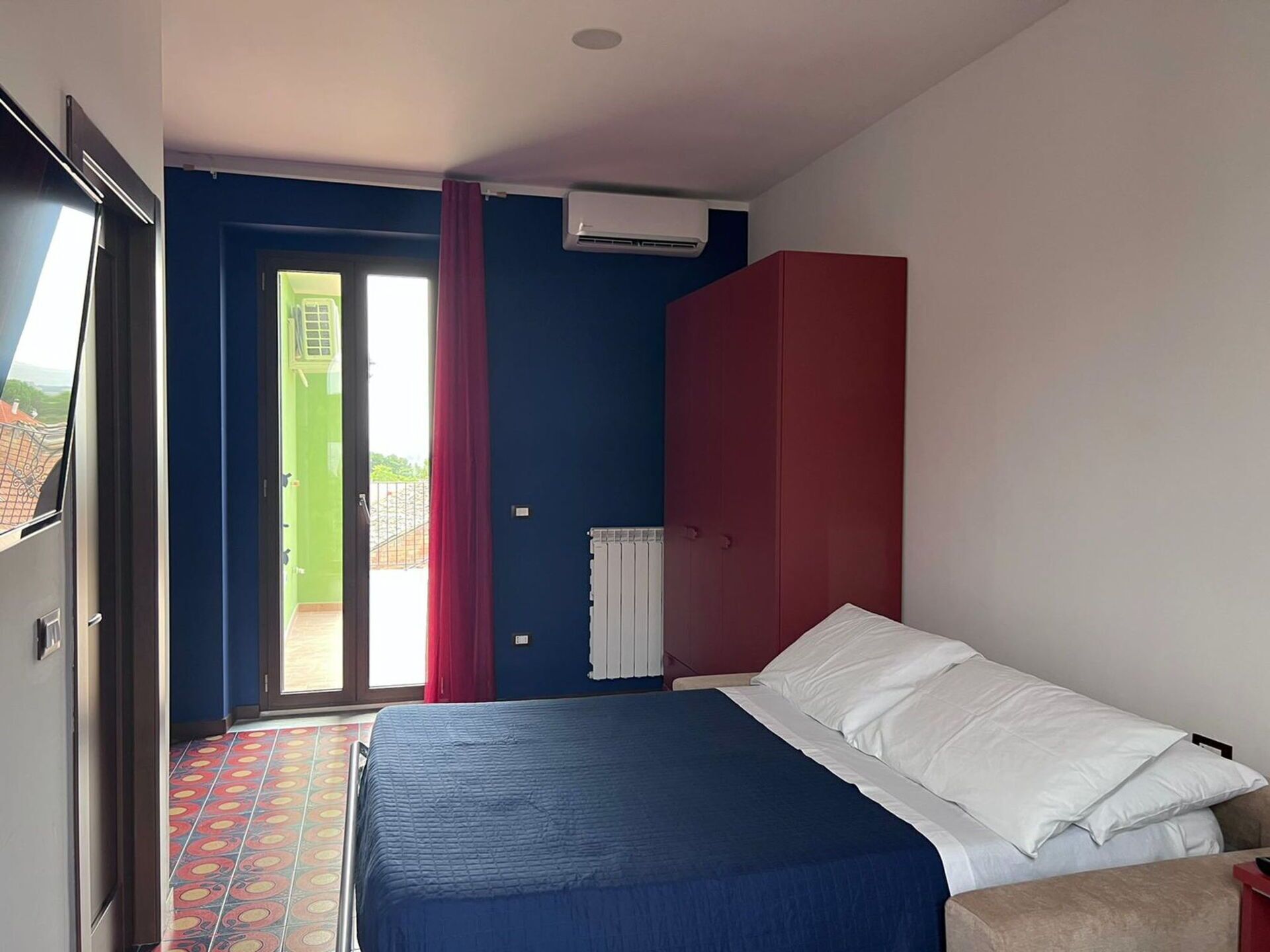 Een slaapkamer met een bed, een rode garderobe en een raam met uitzicht op een groen gebouw.