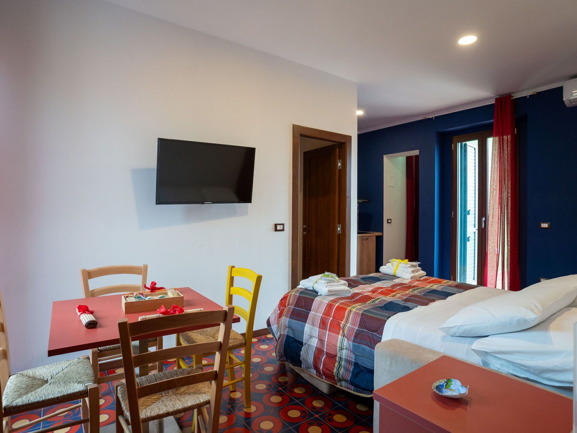 Una habitación de hotel con cama, televisor, mesa de comedor con sillas y una mesita con mantel rojo.