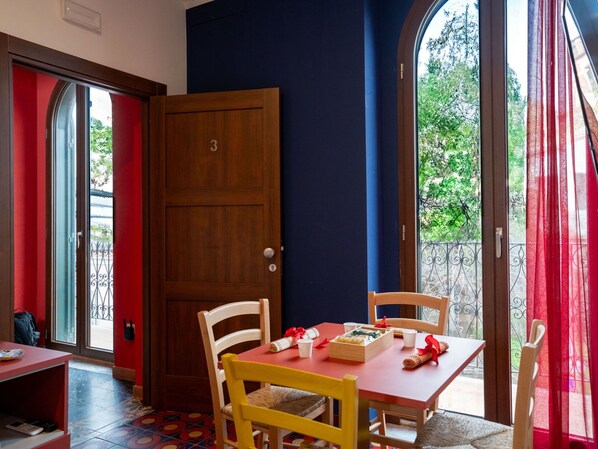 Una habitación con una puerta de madera numerada con el 3, una mesa roja y una silla blanca, y vistas al verdor exterior.