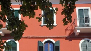 Un bâtiment à la façade rouge, aux ornements blancs et aux volets verts. Il y a deux portes et un balcon avec des balustrades en fer forgé.