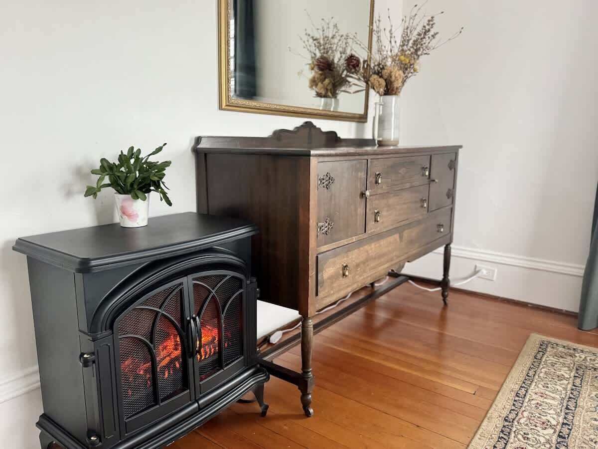 A room with a black wood-burning stove, a wooden dresser, a white chair, and a mirror on the wall.