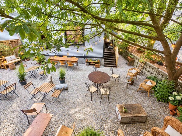 An outdoor patio area with multiple seating arrangements, including tables and chairs, surrounded by potted plants and a tree.