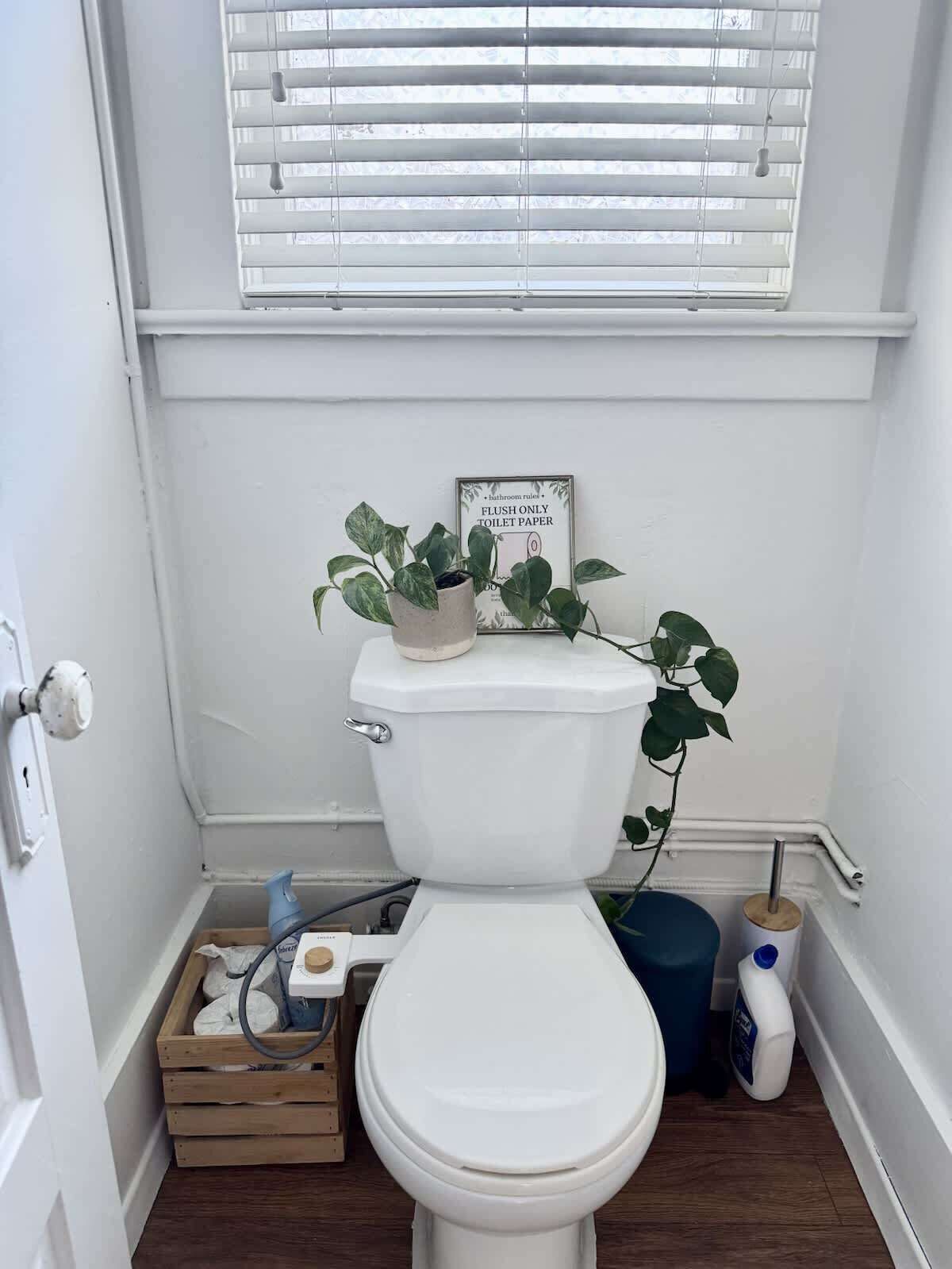 A small, white bathroom with a toilet, a window with blinds, a sign on the wall, and a plant on the toilet tank.
