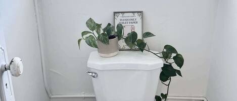 A small, white bathroom with a toilet, a window with blinds, a sign on the wall, and a plant on the toilet tank.