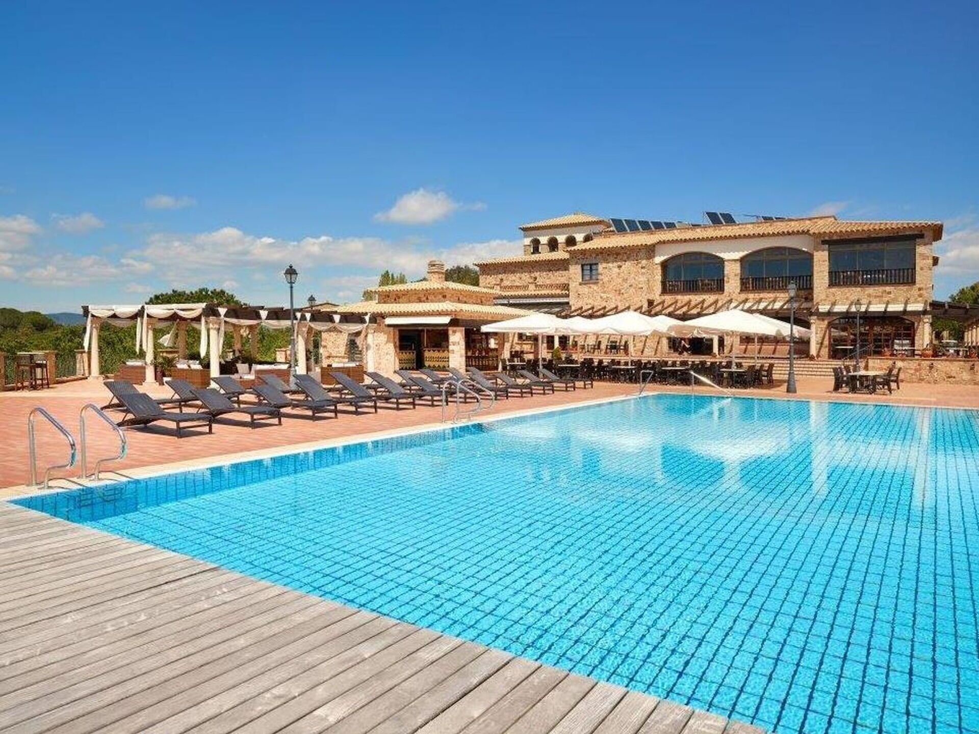 Una piscina con sillones de descanso y sombrillas, un área de estar cubierta y un edificio con tejado de baldosas y grandes ventanales.