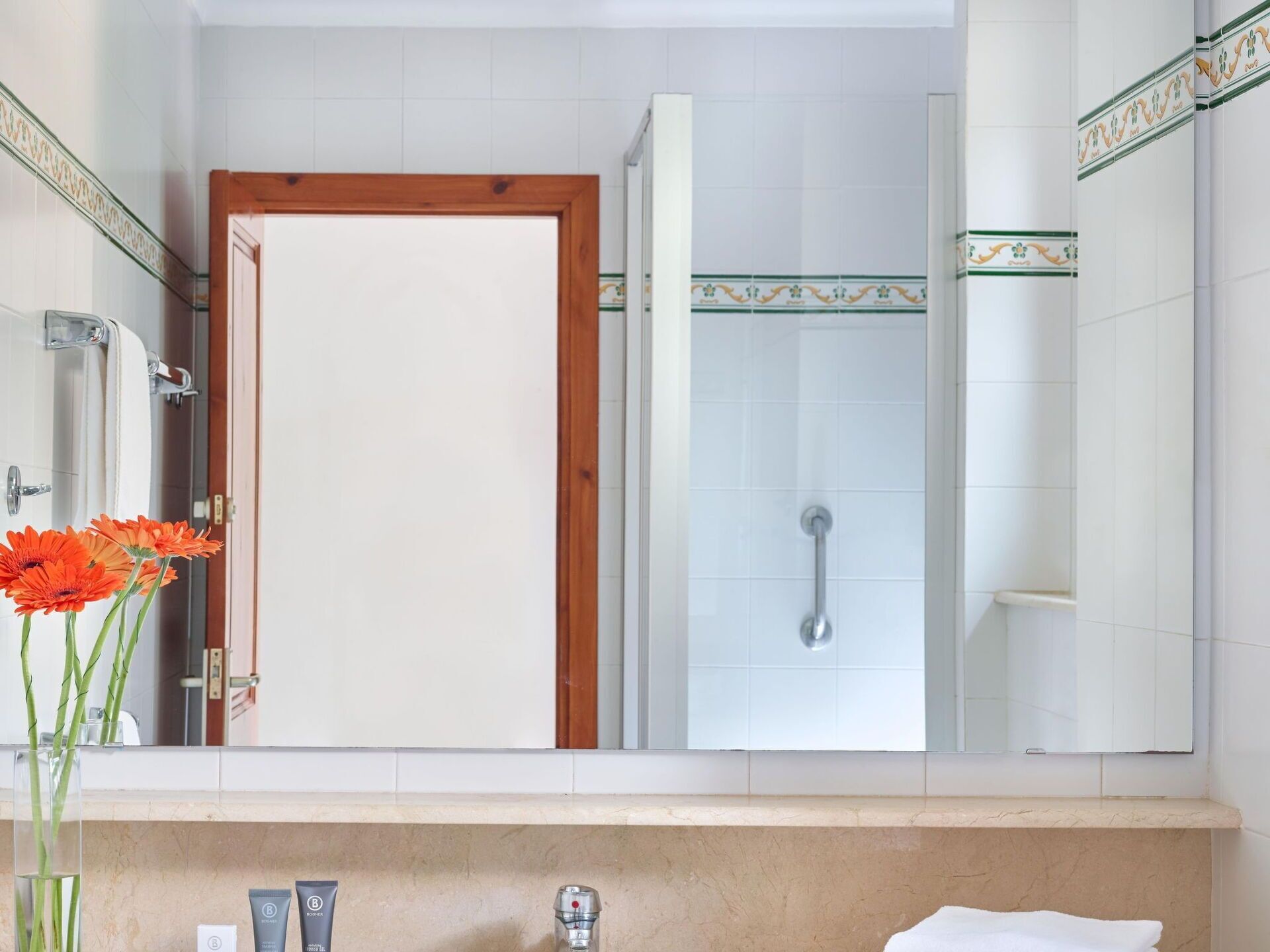 A bathroom with a wooden door, a shower with a handle, a mirror, and a sink with toiletries.