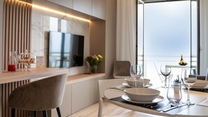 A modern dining area with a wooden table set for two, a television above the counter, and a view of the sea through large windows. - Nice apartment for 3 guests with A/C, WIFI, TV and terrace (Roseto Degli Abruzzi)