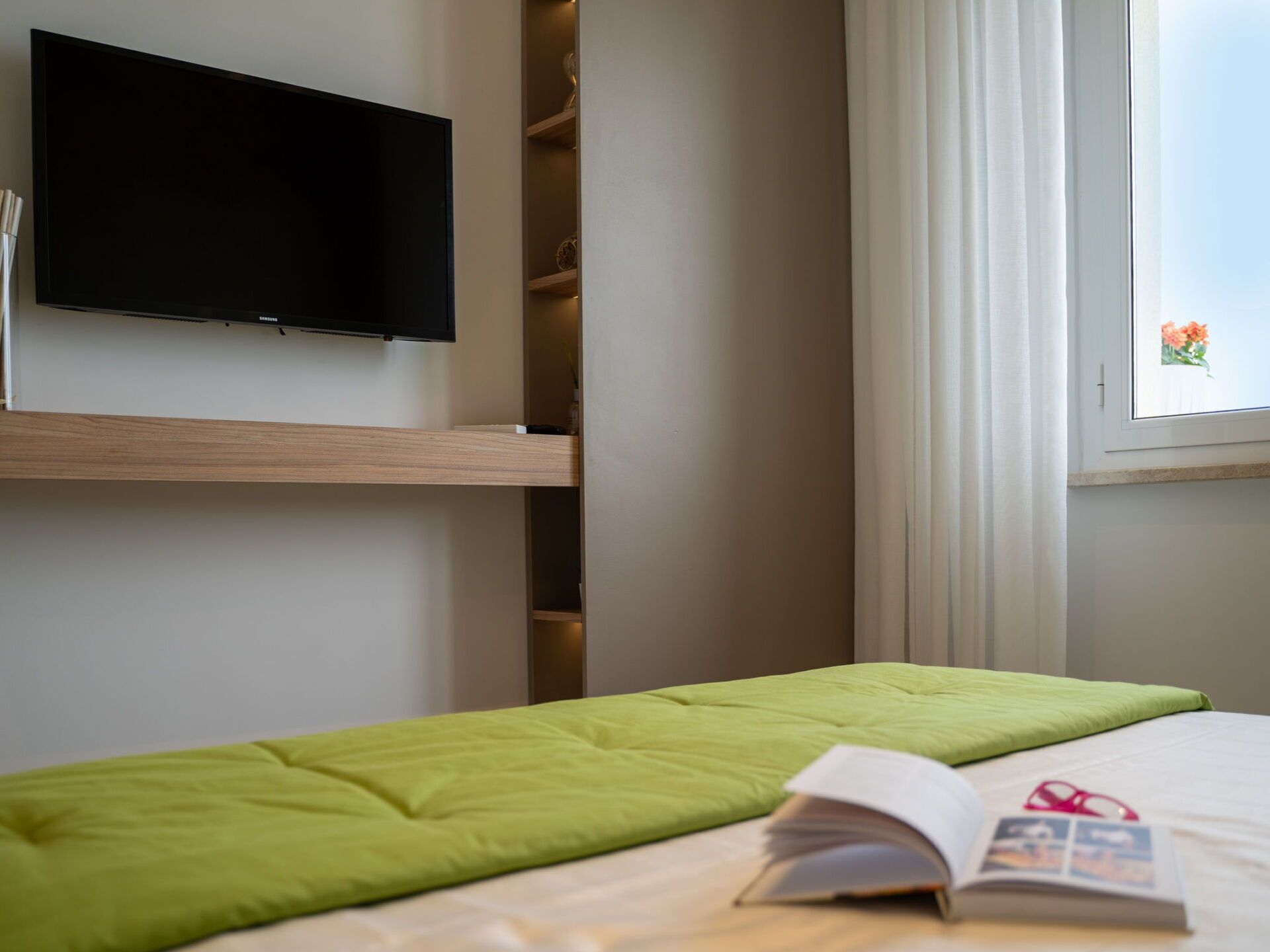 Un dormitorio ordenado con una cama, un televisor de pantalla plana en la pared y una ventana con cortinas.