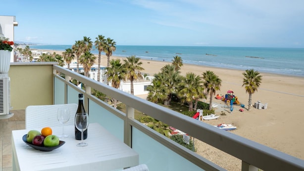 A balcony with a table, a bowl of fruit, and a wine glass. The view includes a beach with palm trees and a clear sky. - Nice apartment for 3 guests with A/C, WIFI, TV and terrace (Roseto Degli Abruzzi)