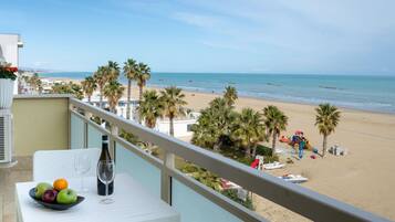 Ein Balkon mit Tisch, Obstschale und Weinglas. Blick auf einen Strand mit Palmen und einem klaren Himmel.