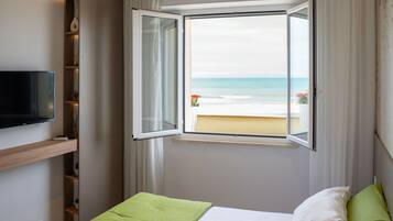 Ein Hotelzimmer mit Bett, einem an der Wand befestigten Fernseher, einem Fenster mit Blick auf den Strand und einem Regal mit Dekoration.