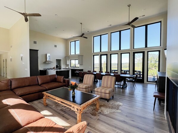 A spacious living area with a large sofa, a coffee table, and a dining table with chairs. There are ceiling fans, large windows, and a kitchen area visible.