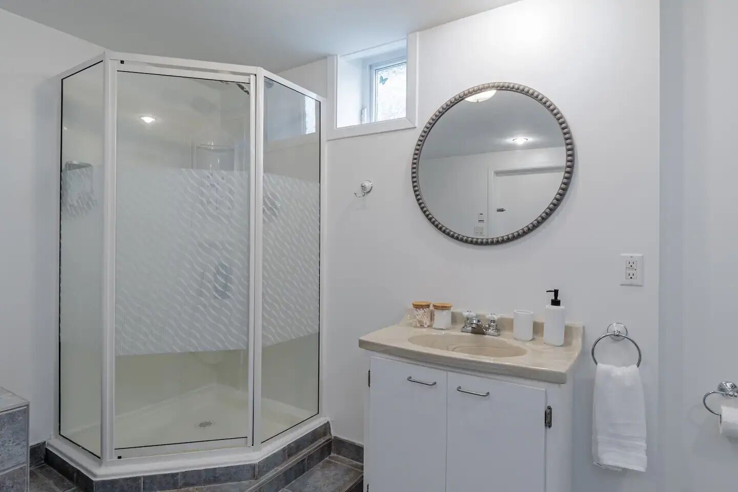 A modern bathroom with a glass-enclosed shower, a round mirror, a sink with toiletries, and a towel ring.