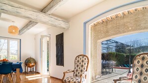 Une pièce spacieuse avec une grande fenêtre, un coin repas avec une nappe bleue et deux fauteuils à motifs.