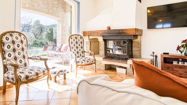 Un salon chaleureux avec une cheminée, deux fauteuils à motifs, un canapé et un téléviseur fixé au mur.