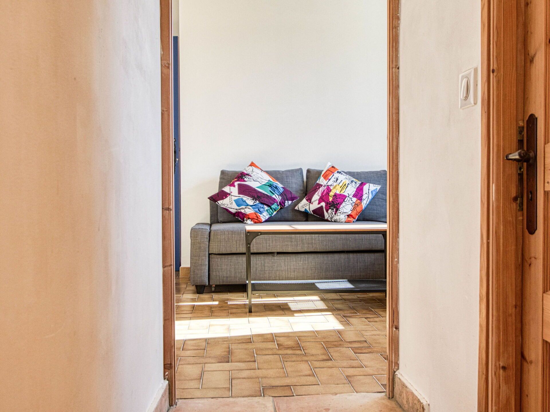 Een gang die leidt naar een kamer met een grijze bank en kleurrijke kussens, een houten deur en een betegelde vloer.
