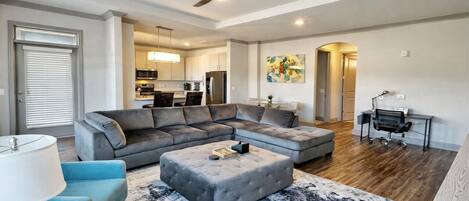 A modern living room with a sectional sofa, a small ottoman, a coffee table, a desk with a chair, and a ceiling fan.