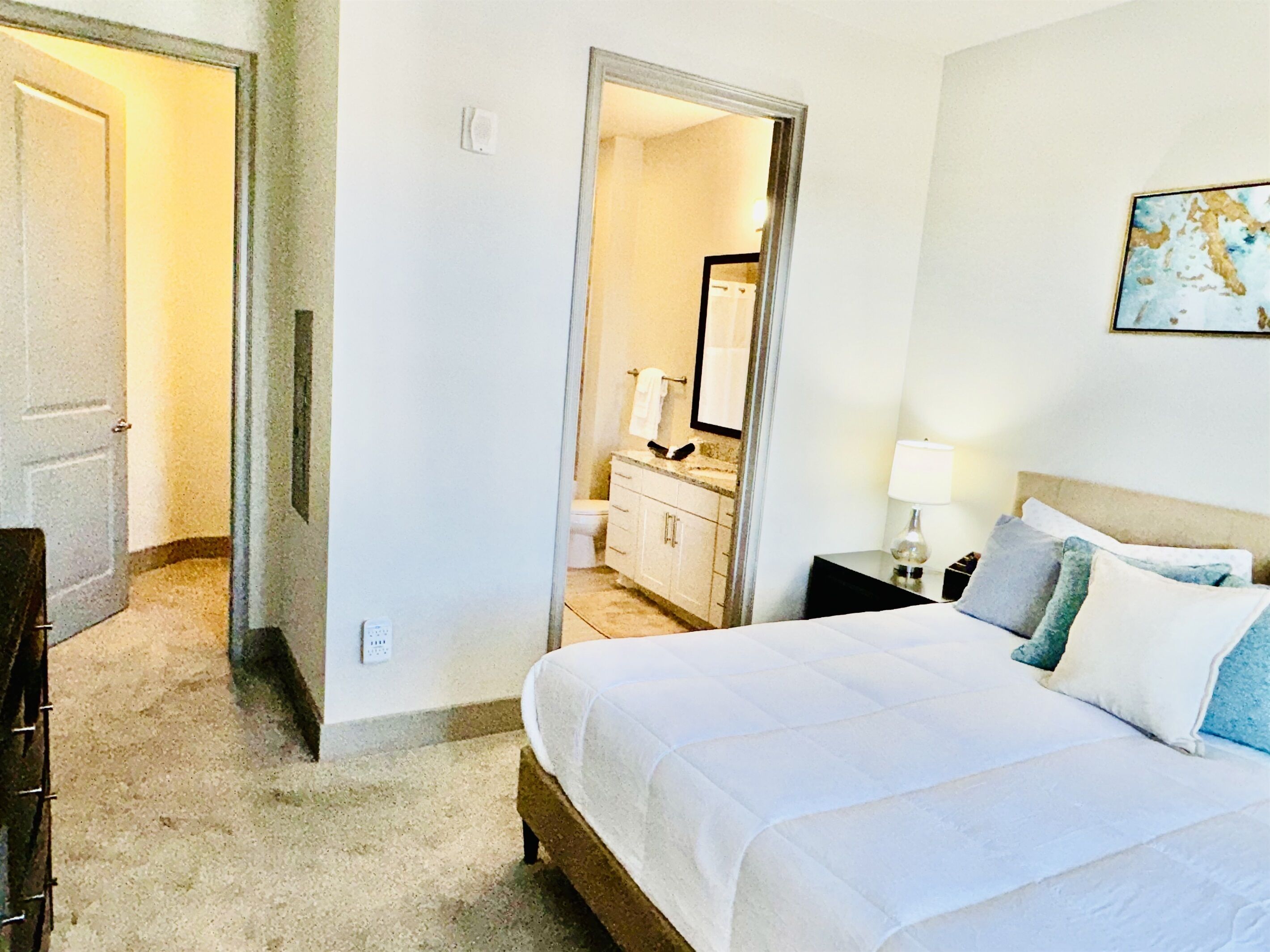 A hotel room with a bed, a nightstand, a lamp, a dresser, and a bathroom visible through an open door.