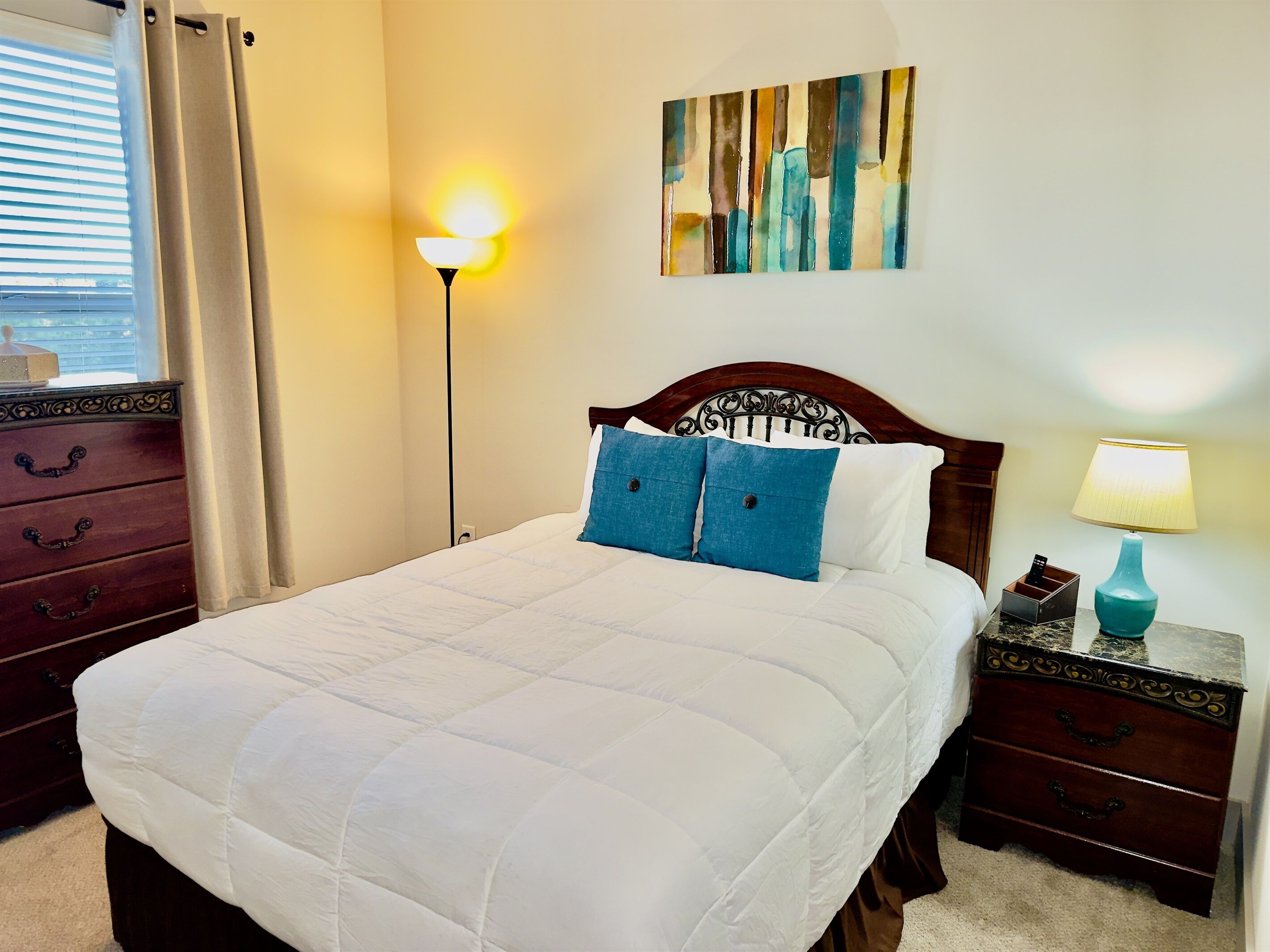 A neatly made bed with a white duvet, two blue pillows, a wooden headboard, a bedside table with a lamp, and a wall-mounted painting.