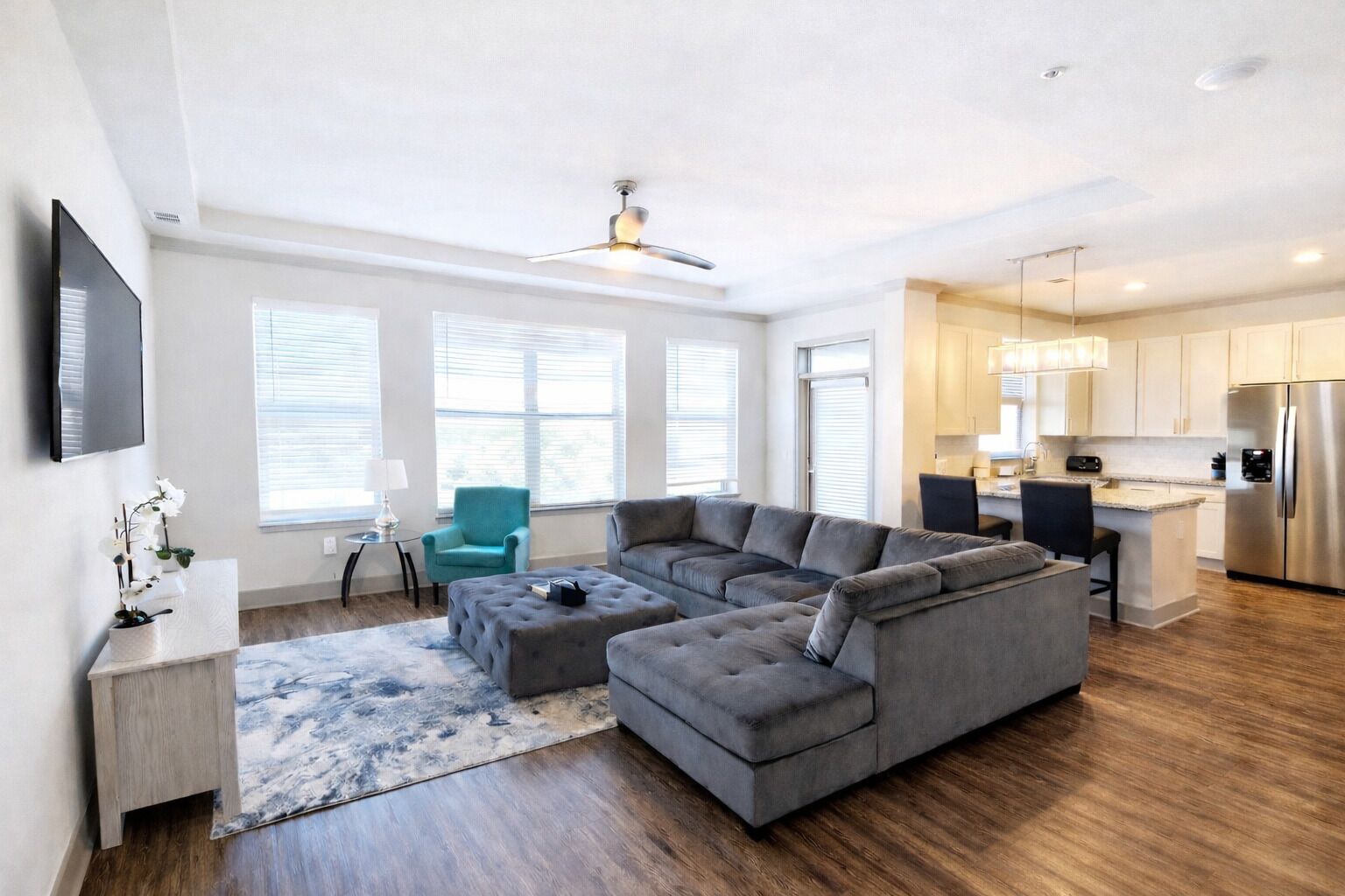 A modern living room with a sectional sofa, a small table, and a TV. There is a kitchen area with a refrigerator and a dining table.
