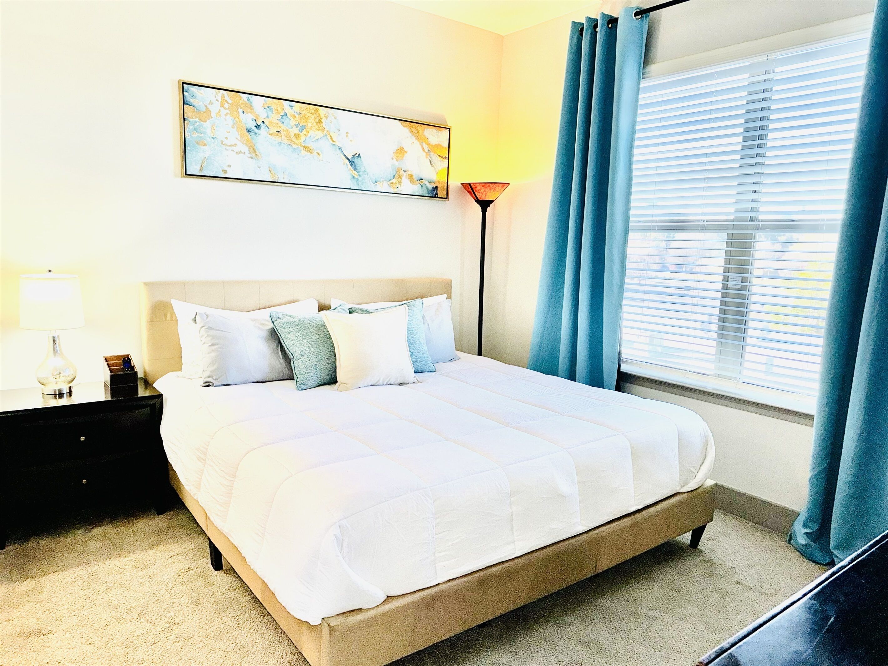 A neatly made bed with a white comforter and pillows, a bedside table with a lamp, a framed abstract painting on the wall, and a window with blue curtains.