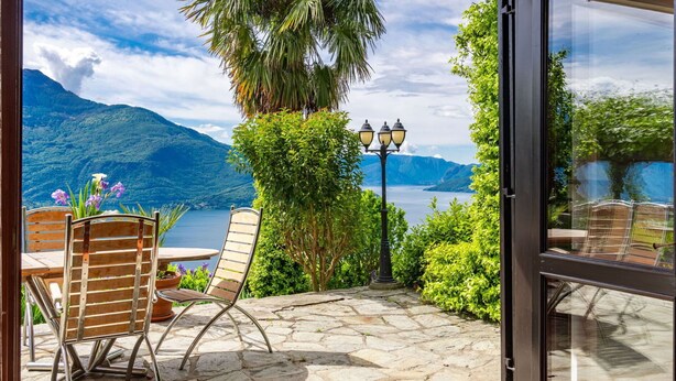 Un patio con pavimento in pietra, due sedie di legno, un tavolo, una lampada e vista su un lago con montagne sullo sfondo.