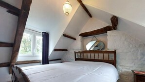 Une chambre avec un plafond en pente, des poutres apparentes, un grand lit, un miroir suspendu et une fenêtre donnant sur les arbres.