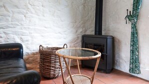 Une pièce avec un canapé en cuir noir, un panier en osier, une table en osier et un poêle à bois noir, le tout placé contre un mur blanc.