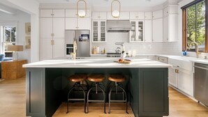 Una cocina moderna con armarios blancos, una isla grande con taburetes, electrodomésticos de acero inoxidable e iluminación colgante.