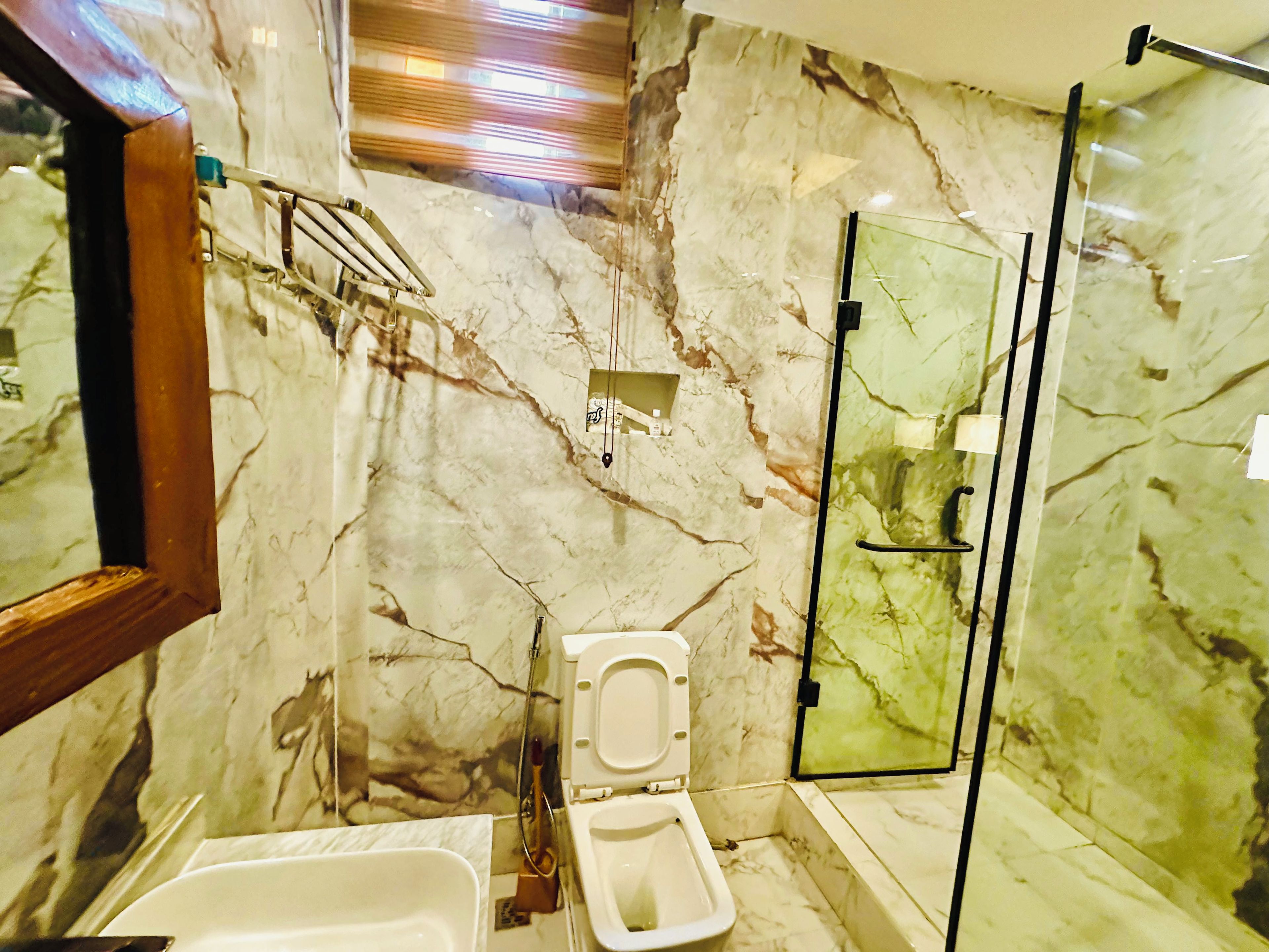 A bathroom with a marble wall, a toilet, a glass shower enclosure, and a wooden-framed mirror.