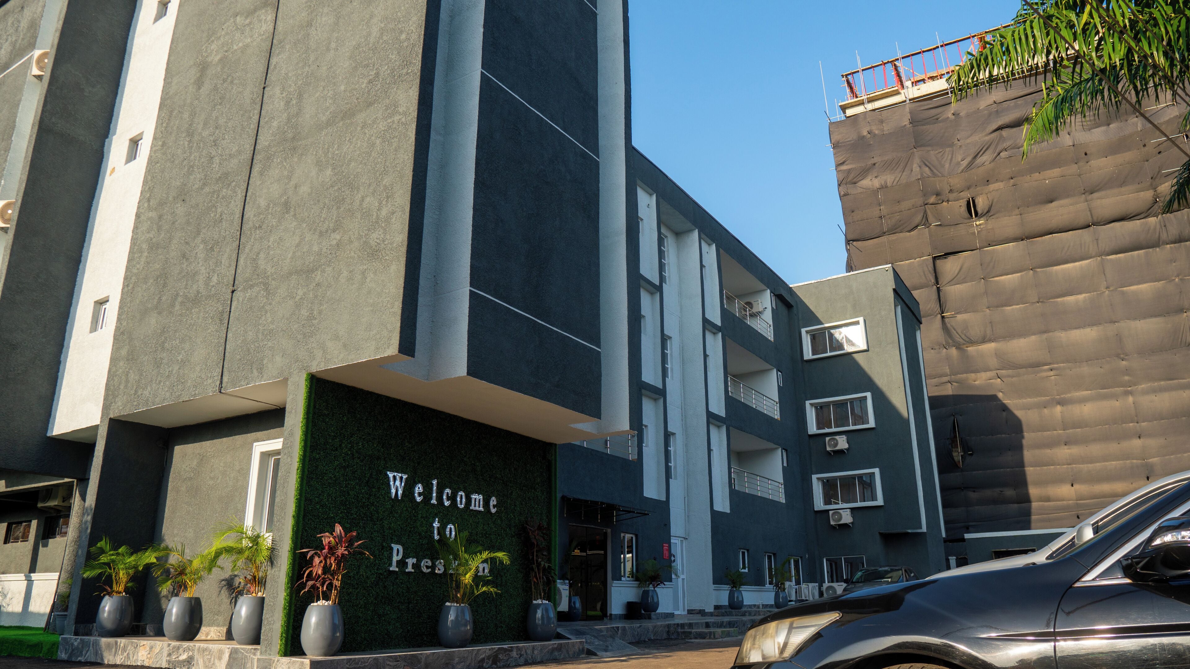 Un edificio moderno con un cartello 'Welcome to Pres', un'auto parcheggiata davanti e un cantiere edile sulla destra.
