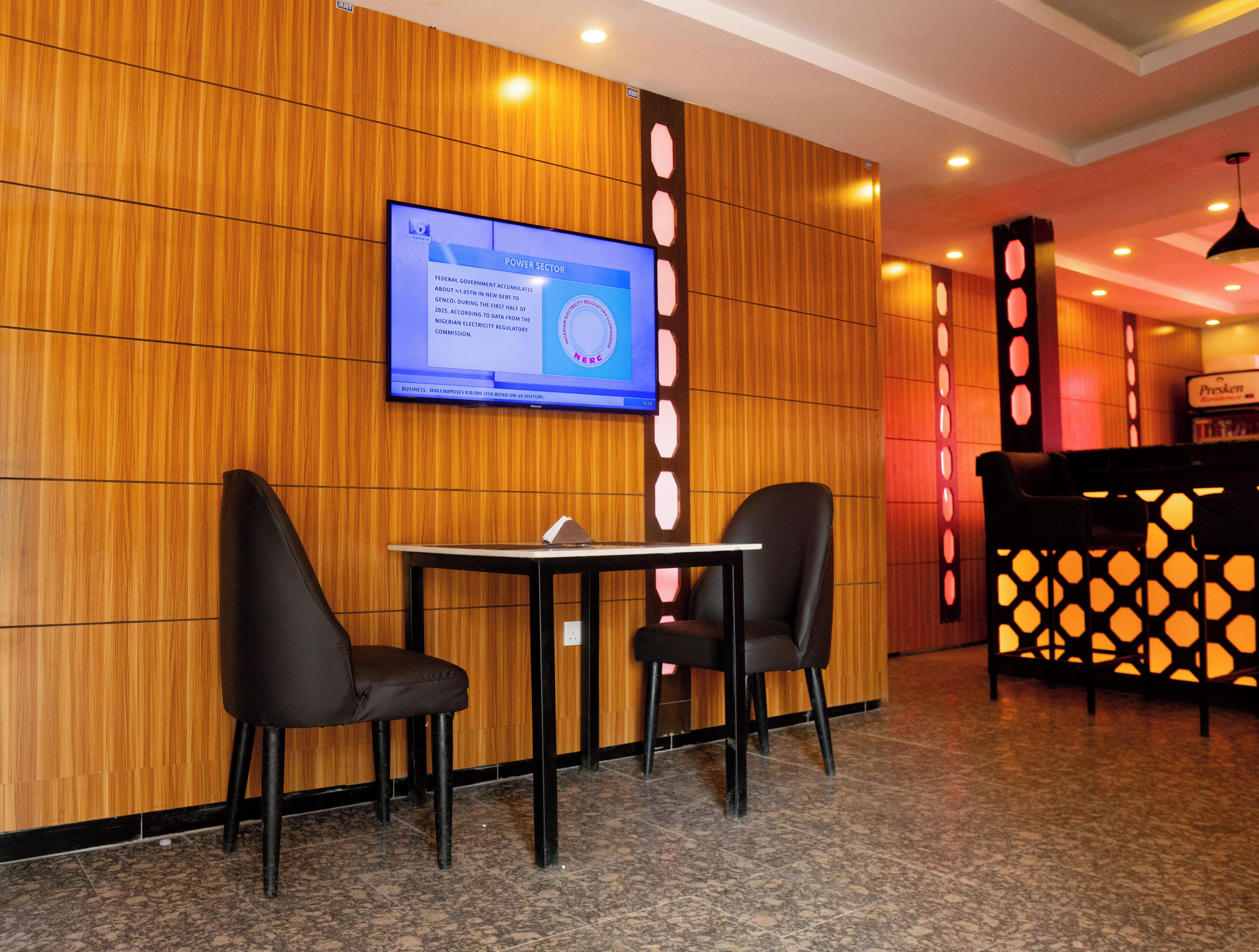 A lobby with a wooden wall, a television screen, two chairs, a table, and a reception desk.