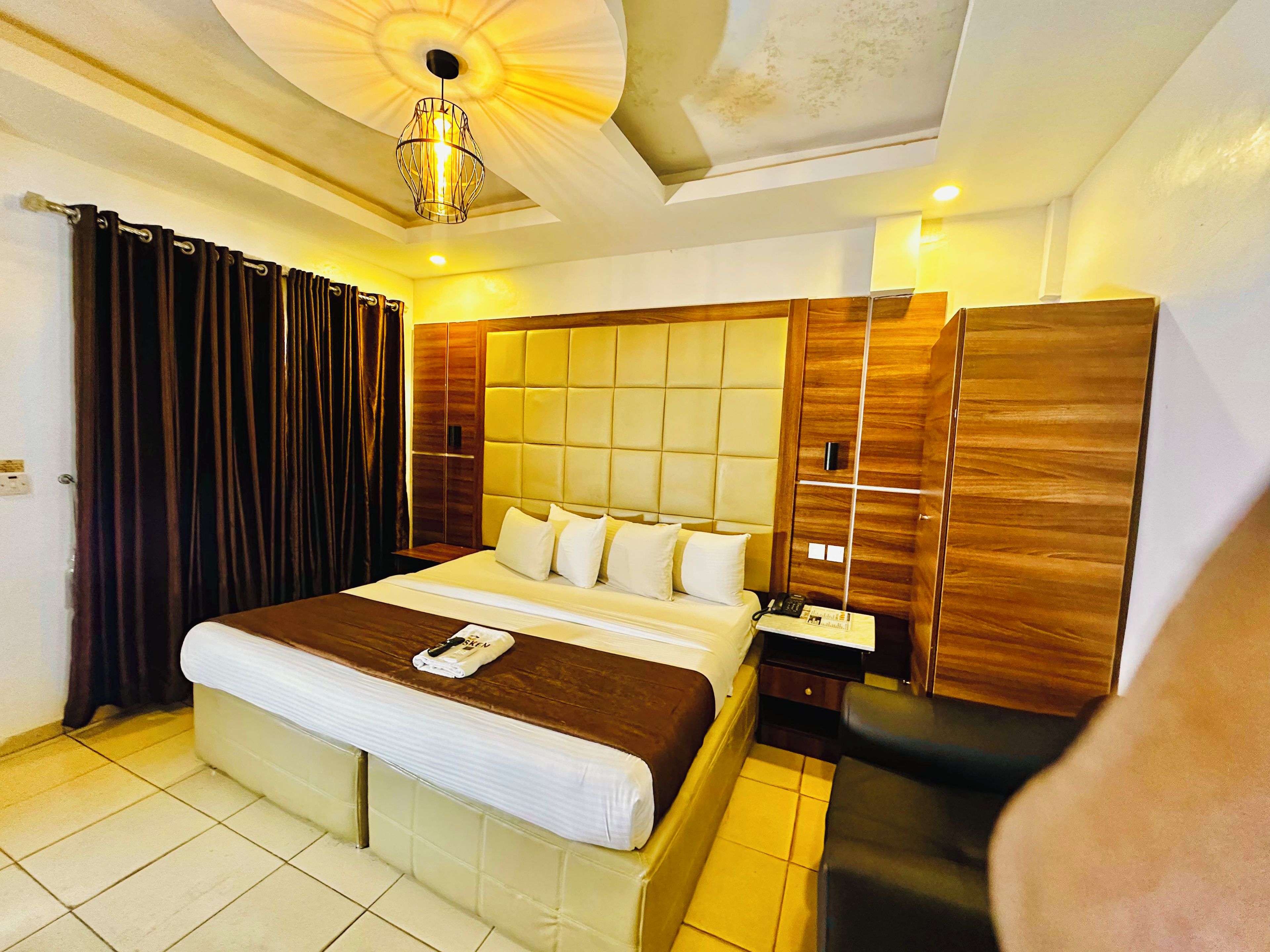 A hotel room with a large bed, a wooden wardrobe, a bedside table with a lamp, and a ceiling light fixture.
