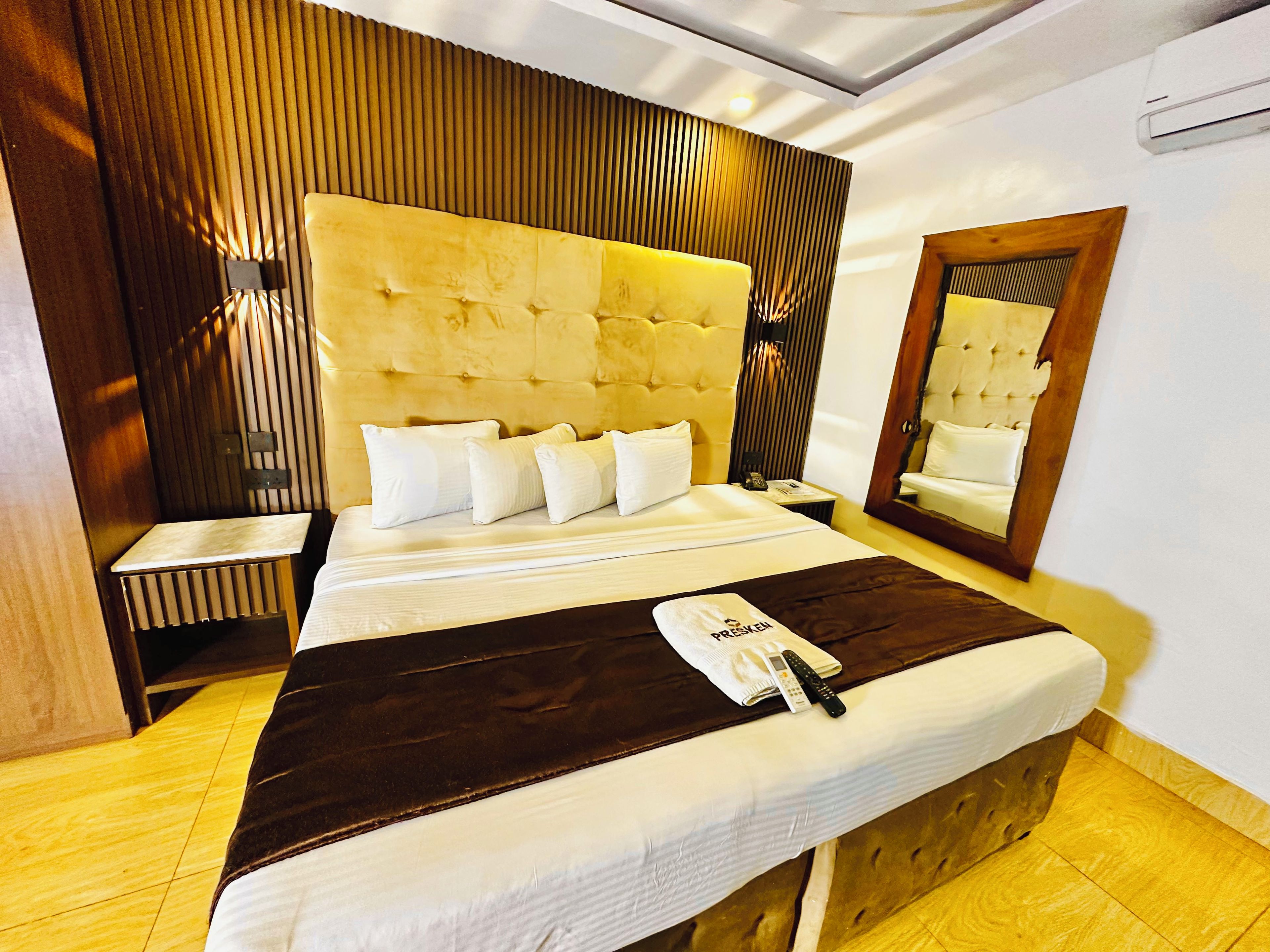 A hotel room with a large bed, a bedside table, a nightstand, a mirror, and a wooden headboard.