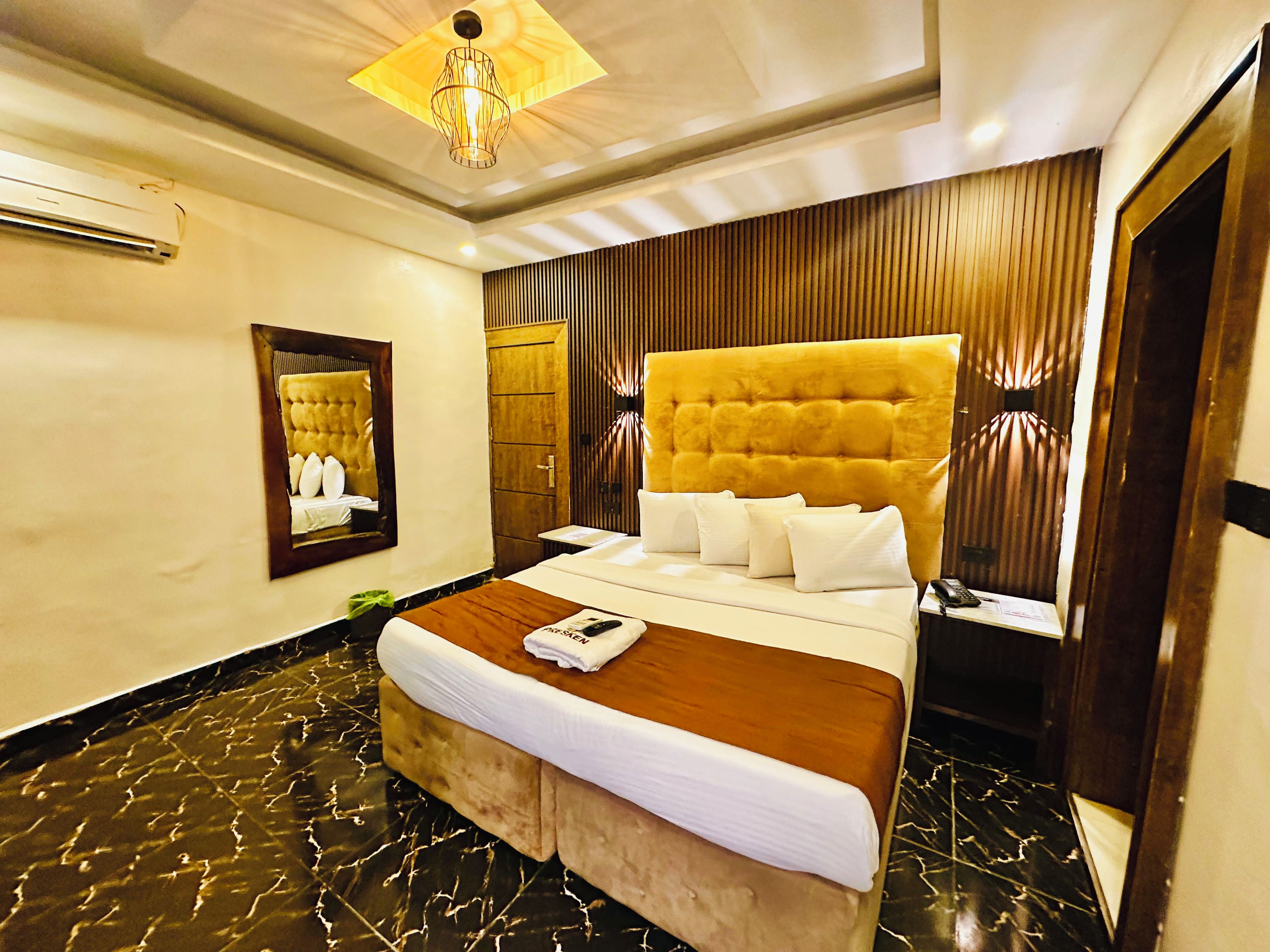 A hotel room with a large bed, a wooden headboard, a mirror, a bedside table, and a modern light fixture.