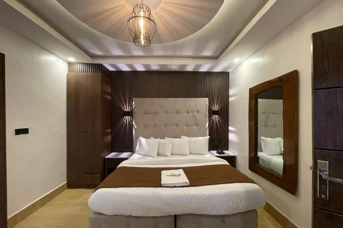 A modern hotel room with a large bed, a wooden wardrobe, a mirror, and a ceiling light fixture.