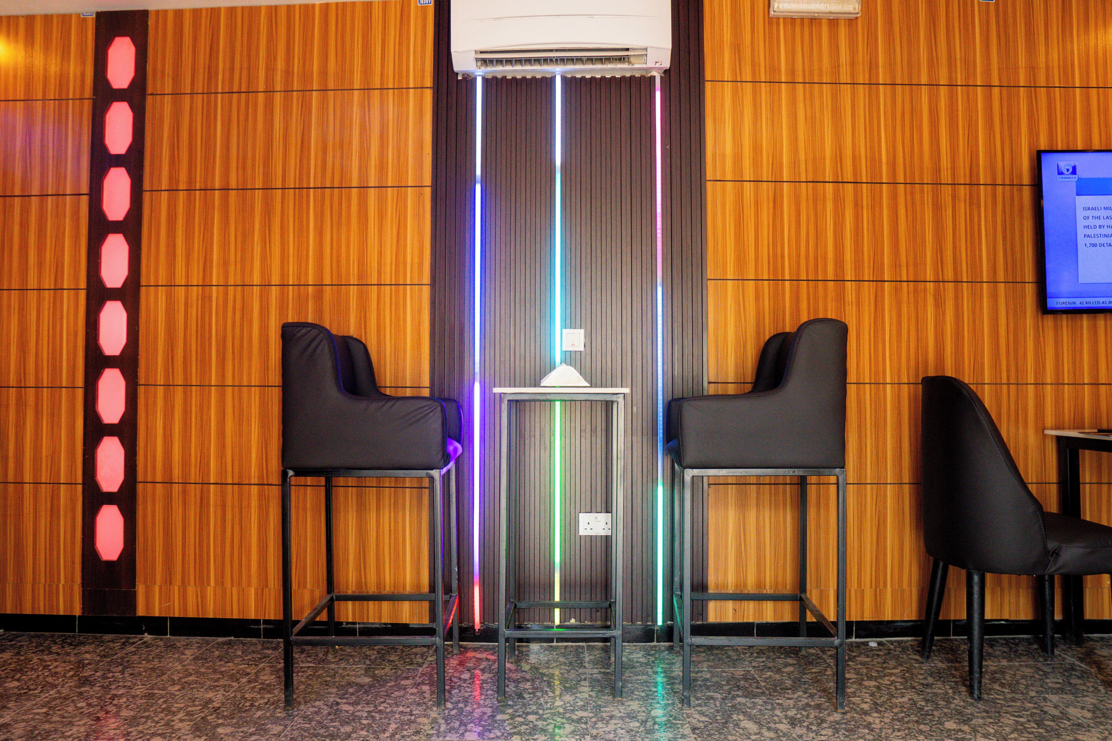 A hotel lobby with wooden paneling, two black chairs, a small table, and a wall-mounted screen.
