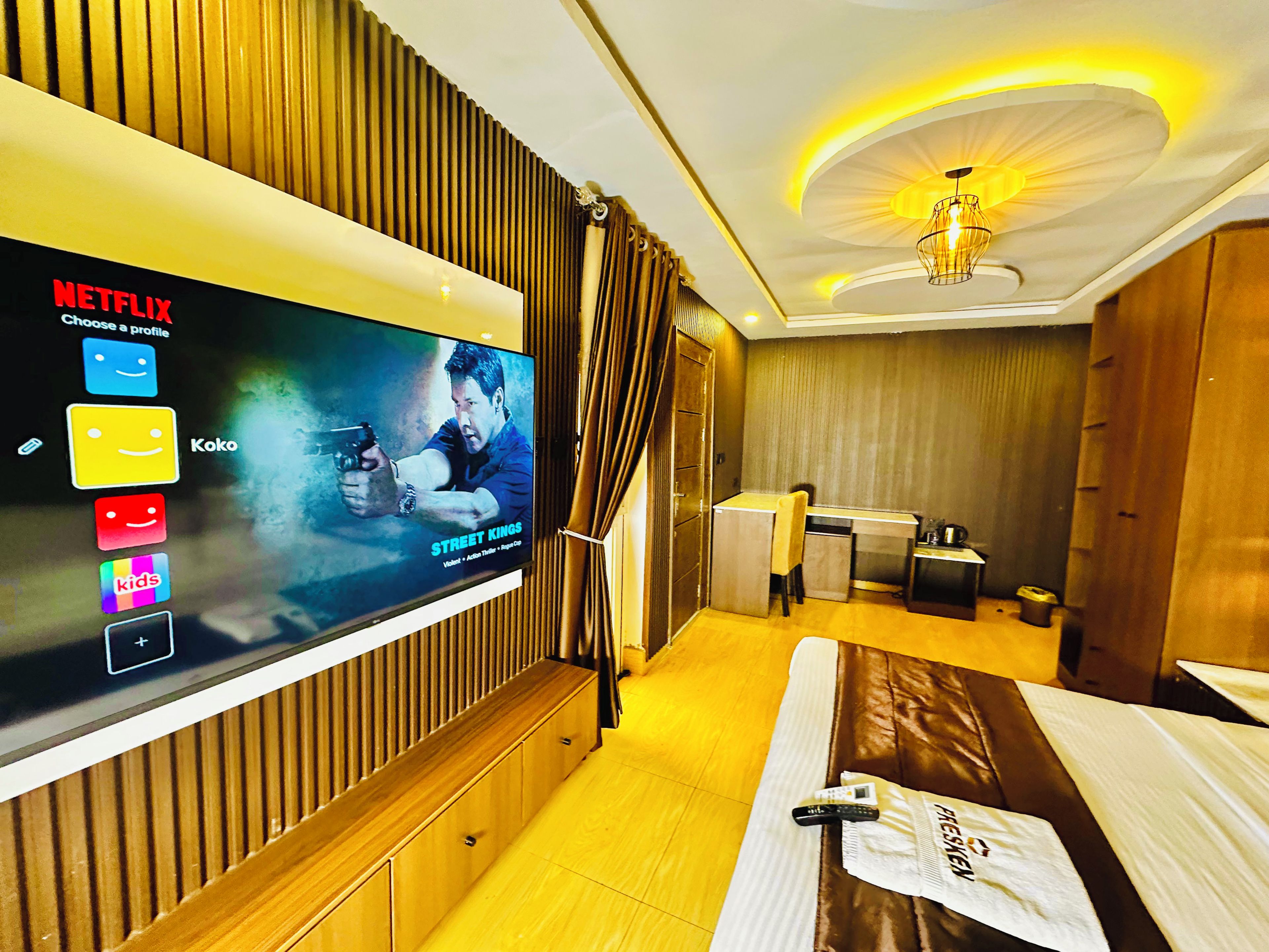 A hotel room with a television displaying a Netflix profile, a desk with a chair, and a bed with a pillow and remote control.